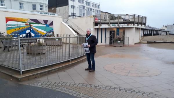 “Person in a dark jacket and jeans holding a newspaper, standing on wet paved ground beside a metal railing in front of a building with a colorful mural and ‘Sandown Hotel’ sign under an overcast sky.”