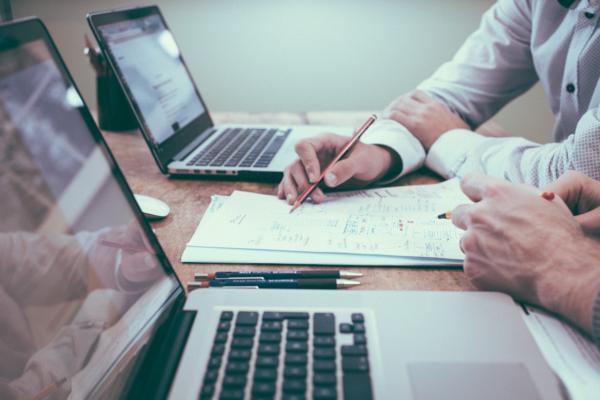 “Two people seated at a desk with open laptops, reviewing and annotating handwritten notes on paper—one pointing with a pencil while the other observes.”