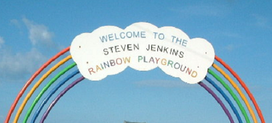 White cloud-shaped sign reading ‘WELCOME TO THE STEVEN JENKINS RAINBOW PLAYGROUND’ mounted above arcs of a rainbow against a clear blue sky.