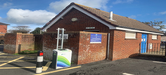 Red-brick public toilet building with a pitched roof and blue door, adjacent to a small electric charging station in a parking area under a clear sky.