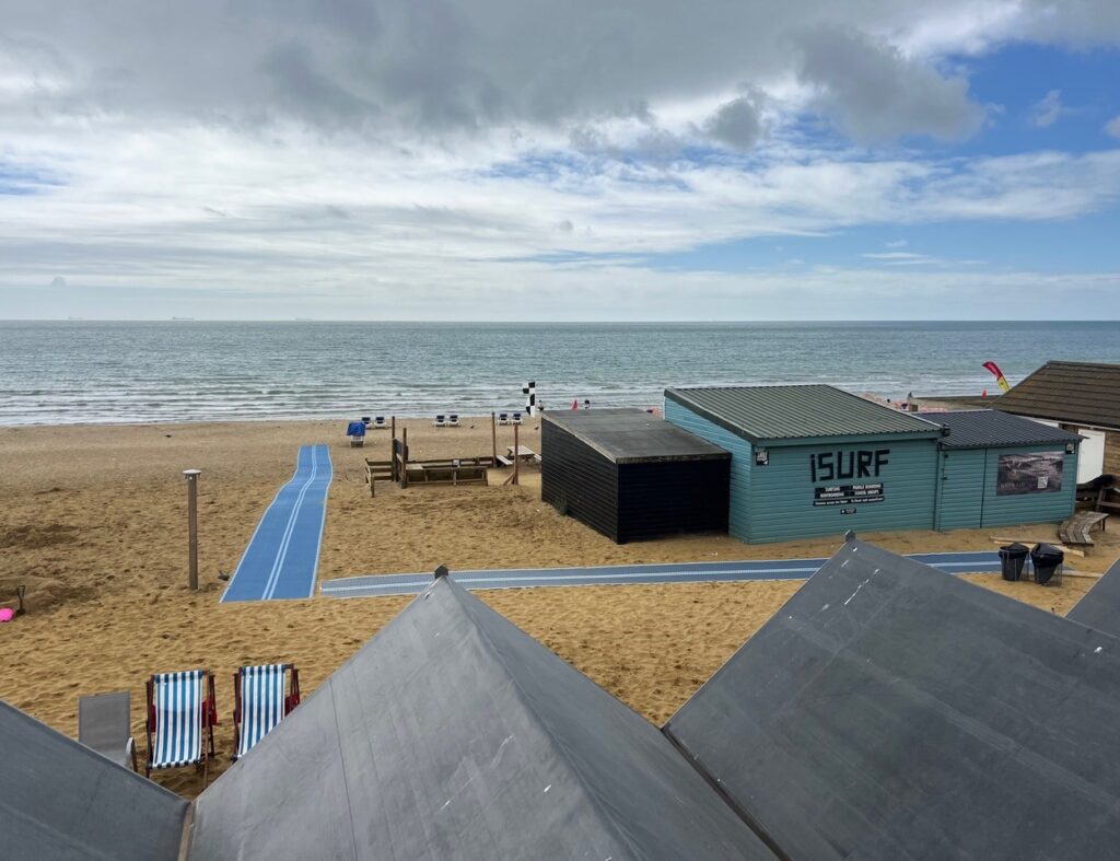 Sandy beach scene with a blue accessible mat leading toward the shoreline; two striped deck chairs sit near the mat’s start.
