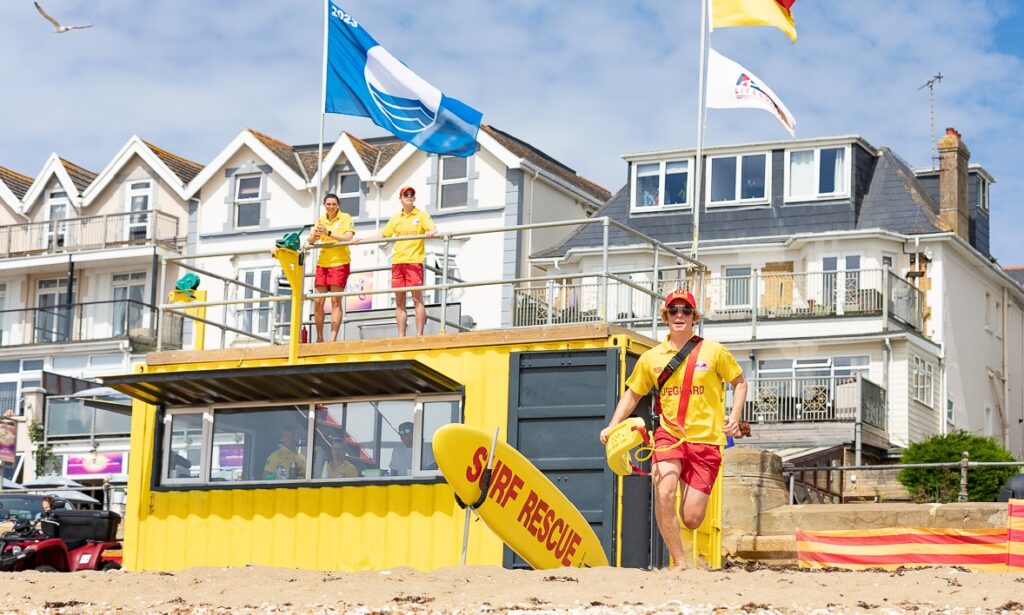 Yellow lifeguard station on a sandy beach with two lifeguards standing on its upper platform.