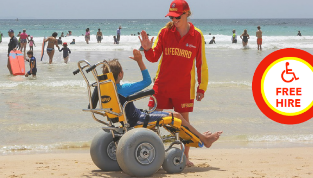 Lifeguard in red uniform high-fiving a person seated in a yellow all-terrain beach wheelchair at the water’s edge.