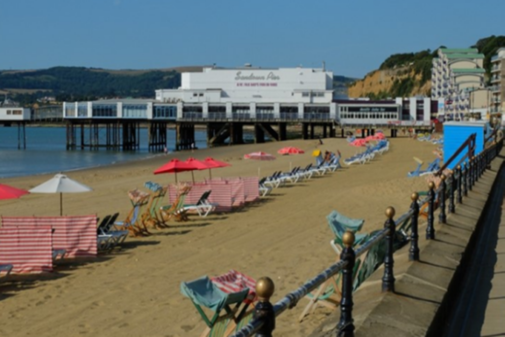 Sandy beach lined with striped windbreaks and rows of deck chairs—green, red, and blue—facing the sea.
