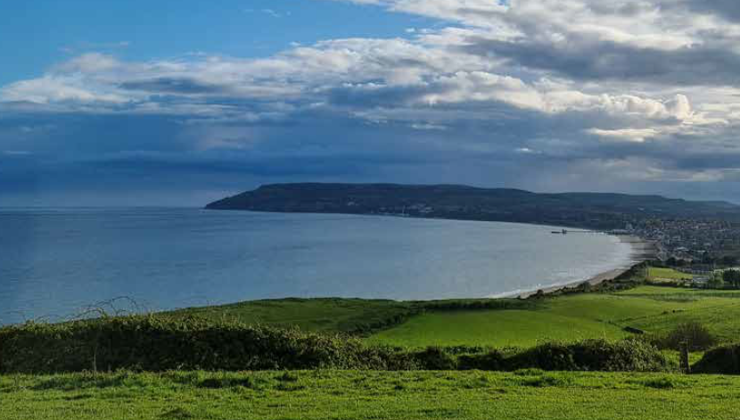 Green fields sloping to a curved bay under a cloudy sky